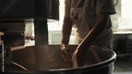 Close up of female baker hands adding yeast or flour into industrial spiral dough mixer bowl for kneading process. Production of fresh baked goods in professional kitchen. Female baker adds yeast to b