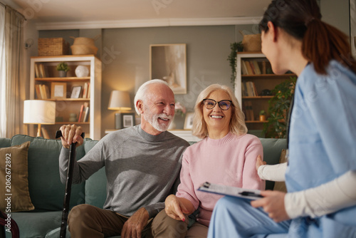 A home health nurse meets with an elderly couple in their living room. The nurse talks with them while they sit together and smile. The setting is bright and welcoming.
