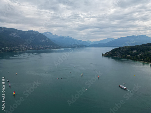 Aerial Drone View of Lake Annecy with Sailboats, Windsurfers, and Passenger Boat Surrounded by Mountains Under Cloudy Sky, Haute‑Savoie, French Alps, France