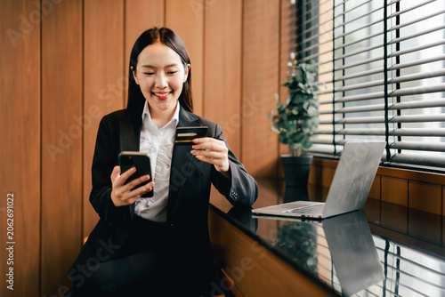 Asian woman making online payment with credit card and smartphone, engaging in digital banking, e-commerce, and secure mobile shopping at a modern workspace