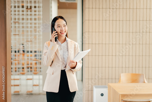 Young asian businesswoman smiling on a phone call while walking through a bright minimalist office, holding documents — confident, productive and connected in a modern workplace