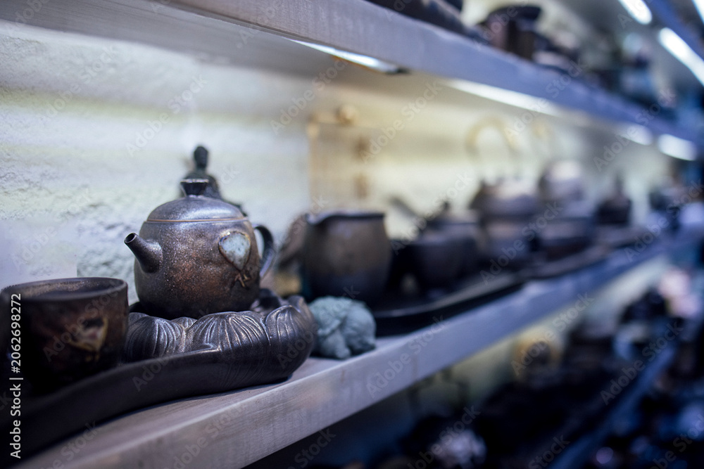 Fototapeta premium Traditional ceramic teapots and cups displayed on a wooden shelf in a tea shop, showcasing various shapes and sizes with a soft, illuminated background