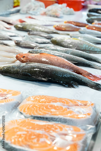 A traditional Taiwanese fish market stall showcases fresh seafood displayed on crushed ice, including whole snappers, dark-scaled fish, and vibrant salmon slices. The composition highlights freshness,
