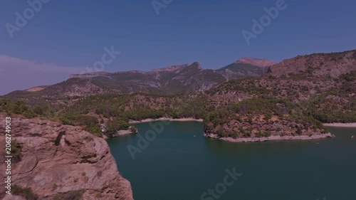 Drone footage blasting past a rugged rock outcrop to suddenly expose a secluded teal cove, pine-fringed shoreline, and a dramatic limestone mountain range under vivid blue sky.