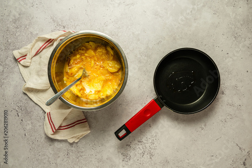 Vista superior de un bol con patatas y huevo batido, junto a una sartén para preparar una tortilla de patatas