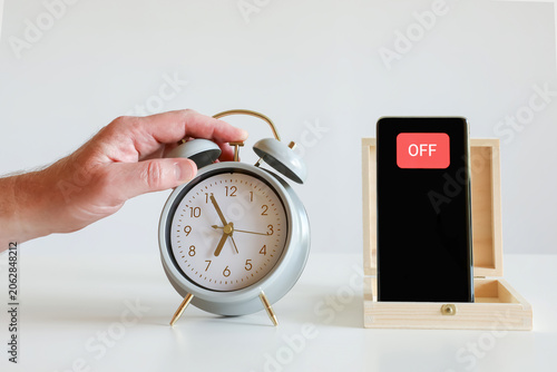 Human hand turning off alarm clock next to smartphone in wooden box. Digital detox, screen time reduction, offline lifestyle, slow living, technology break, mindful living concept.