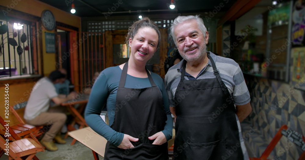Obraz premium Young woman and senior man small business workers standing together inside café wearing aprons smiling at camera expressing teamwork pride and daily hospitality work
