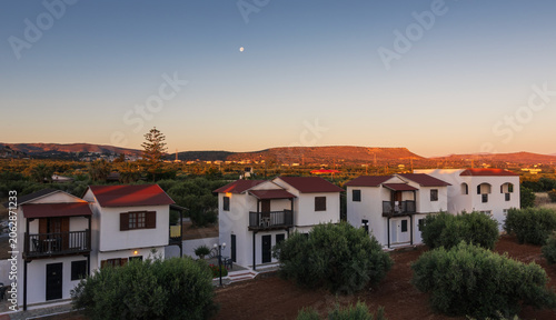 Scenic sunset landscape in Crete with white holiday villas, green trees and mountain ridges in the background during a golden hour. Kato Gouves, Greece island