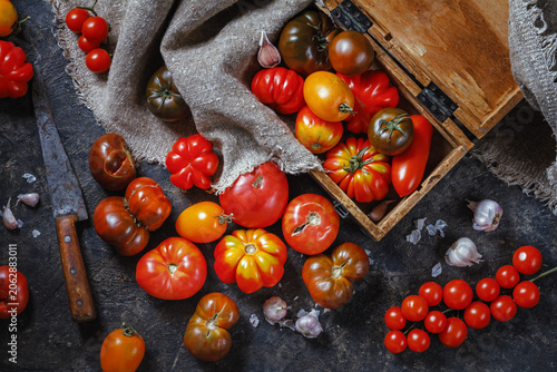 Photos Many different breeds, shapes and sizes of tomatoes in an old wooden box and on a dark surface, flat lay, top view