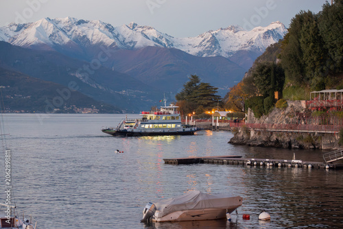 A tranquil scene unfolds at dusk on a lake, with a ferry approaching a shore dotted with lights, framed by majestic snow-capped mountains in the background