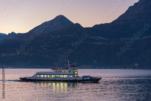 A ferry boat glides across a tranquil lake at dusk, silhouetted against a backdrop of majestic, dimly lit mountains