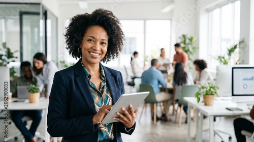 Confident African American businesswoman with tablet smiling in modern open plan office with diverse team working in background commercial corporate professional workspace leadership executive
