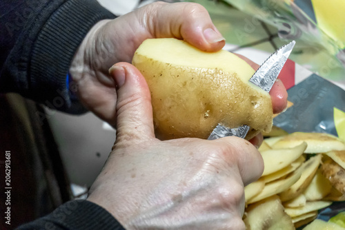 Primer plano de manos de mujer mayo pelando una patata con un cuchillo de cocina pequeño, mostrando las peladuras en el fondo durante el proceso de preparación de ingredientes caseros tradicionales.