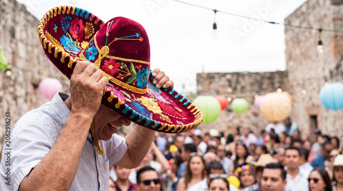 Man wearing decorated sombrero at Mexican holiday celebration. Joyful latino event in courtyard with lantern. Traditional cultural festival party with crowd of people gathering together.