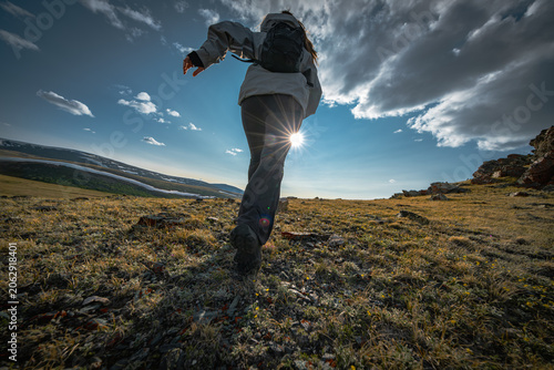 Low angle rear view of active woman with backpack running up rocky mountain hill. Female tourist exploring remote wilderness under dramatic sky with bright sunburst flare.