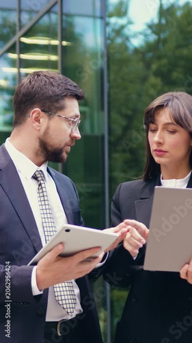 Two business colleagues, man and woman, talking and working on a laptop and tablet while walking outdoors near a modern office building. Slow motion