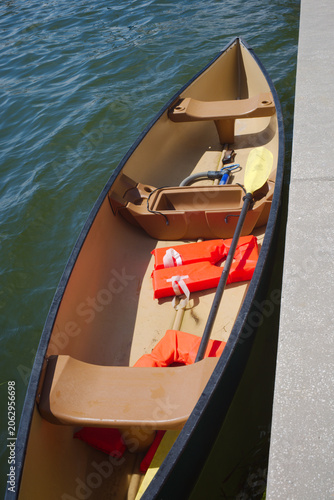 A tan canoe tied to a concrete dock holds bright orange life jackets and a yellow paddle, floating on gentle rippling lake water under clear sunny light for a relaxed outdoor day.
