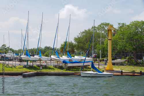 Schilderij op canvas Sailboats with blue covers lined up on trailers beside a marina dock, waterfront water in Gulfport, FL the foreground and trees in the background conveying leisure, calm and summer boating lifestyle