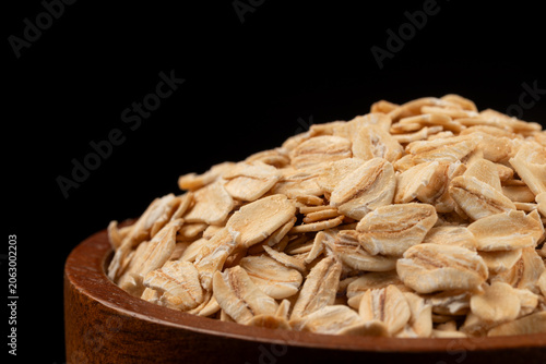 Pile of raw whole grain rolled oats in wooden bowl on black background.