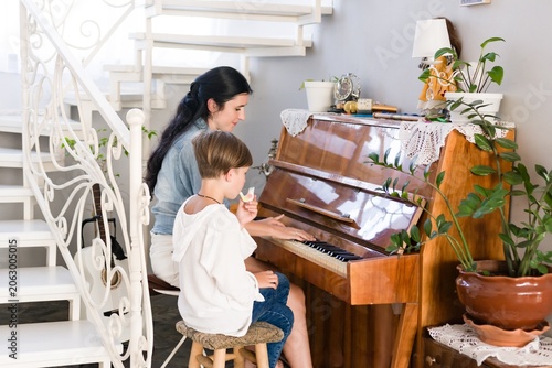 A mother and child share a moment of musical learning. The child sits at an upright piano, receiving instruction from the woman. The setting is a home with stairs and plants visible.