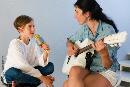A boy holds a microphone, singing along to a woman playing a white acoustic guitar. They are sitting close, sharing a musical experience in a bright, casual setting.