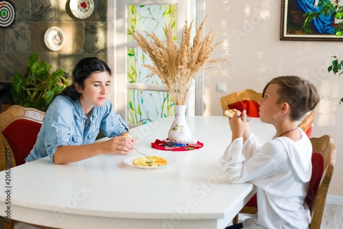 A woman and a young boy are seated at a white dining table, each enjoying their food. The scene is peaceful and intimate, set in a sunlit room with decorative elements.