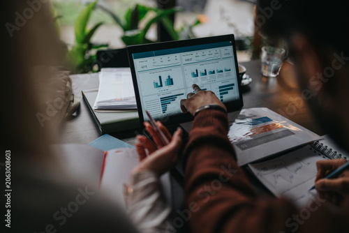 Two business colleagues analyze charts and data on a laptop screen while taking notes. The scene shows a collaborative meeting focused on analytics and planning.