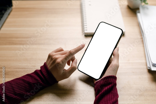 Hands of a woman holding and pointing at a smartphone with a blank white screen, sitting at a wooden desk in an office workspace