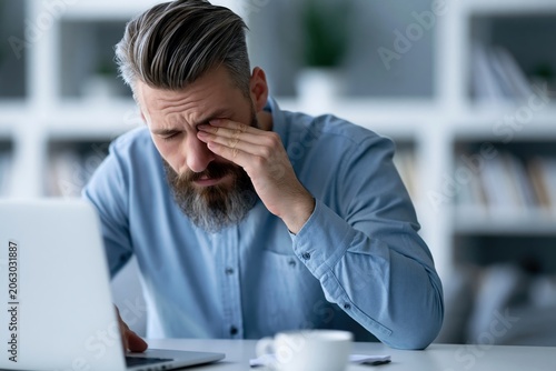 Man rubs his eyes while working on a computer at home during the day in a modern office setting