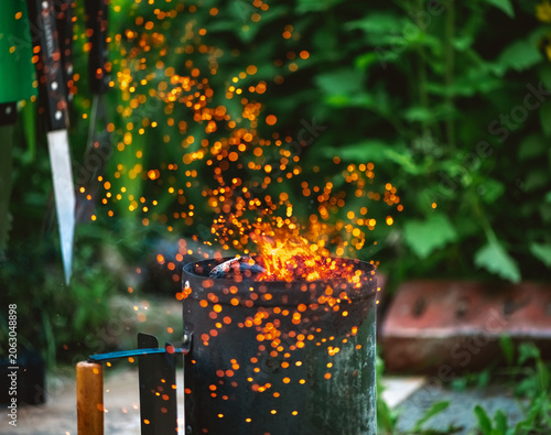 Charcoal briquettes are igniting inside a metal chimney starter, generating brilliant orange sparks and flames, preparing for outdoor grilling and cooking in a garden setting