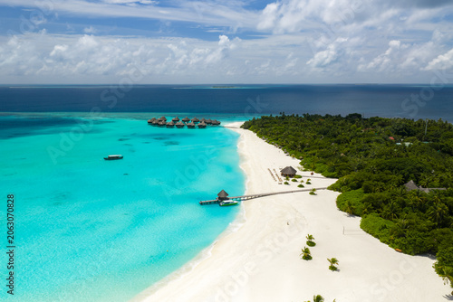 Foto Aerial Maldives island resort shows white sand curving by turquoise shallows, overwater villas on stilts, a wooden jetty, thatched pavilions, and the deep Indian Ocean