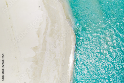 Aerial view of Maldives shoreline shows powder white sand meeting clear turquoise water. Gentle waves shimmer in bright daylight, with sandbars visible and no people present.