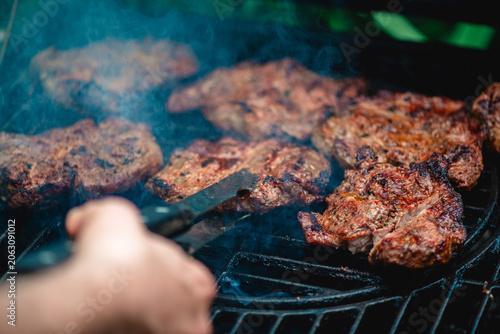 Canvastavla Hand holding tongs turning multiple cuts of marinated meat sizzling on a hot gri