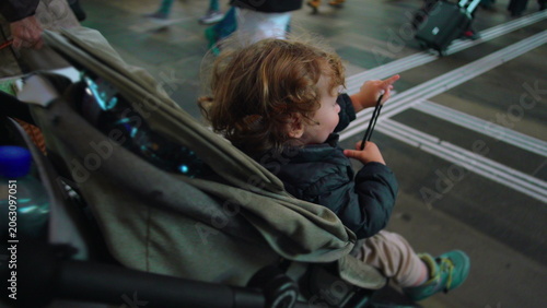 Toddler seated in stroller at busy station looking upward with curiosity as travelers pass by evoking wonder transition public travel movement and early social awareness