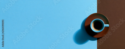 A single cup of coffee in a saucer on a blue and brown surface with shadows