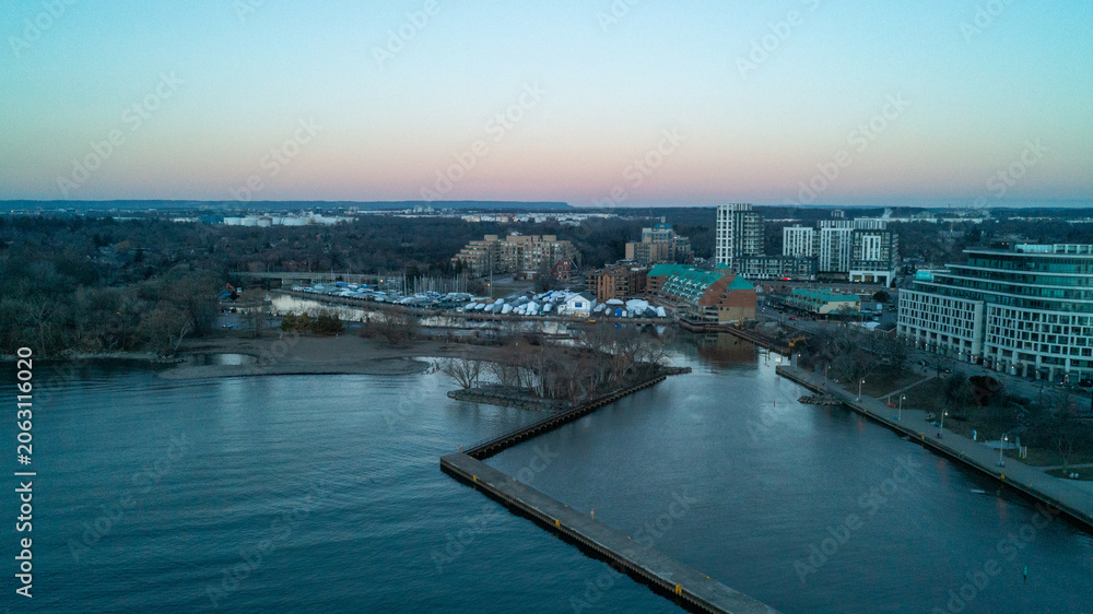 Naklejka premium A beautiful high-angle drone photograph over Lake Ontario, capturing the long L-shaped concrete pier at Bronte Harbour, empty winter boat slips, and waterfront buildings under a twilight sky.