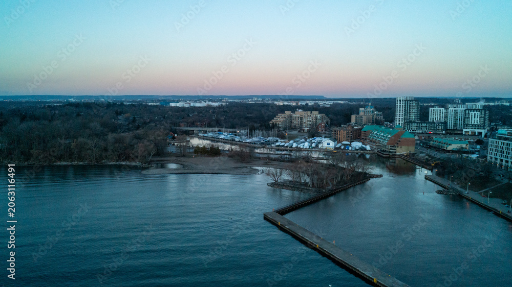 Obraz premium A beautiful high-angle drone photograph over Lake Ontario, capturing the long L-shaped concrete pier at Bronte Harbour, empty winter boat slips, and waterfront buildings under a twilight sky.