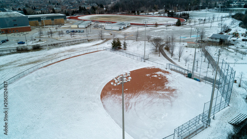 Fotografia A crisp high-angle shot capturing a snow-covered baseball field with its brown infield exposed, overlooking a high school campus, running track, and suburban park on an overcast winter day