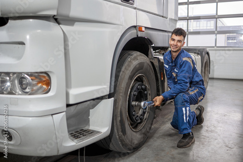 A smiling mechanic expertly uses a professional tool to unscrew a wheel from a truck in a well-lit workshop. The image captures his concentration and skill, showcasing automotive care.