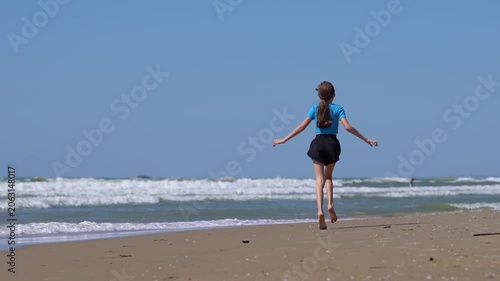A teenage girl strolls along a beach with waves. A girl walks along the seashore or ocean on a sunny day.