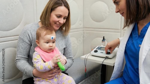 Female audiologist placing an electrode on a baby's forehead for an auditory brainstem response test, while the infant's mother holds her child during the medical examination in a clinic