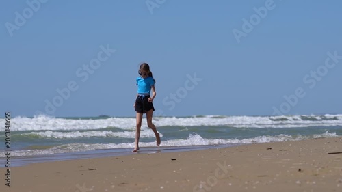 A teenage girl strolls along a beach with waves. A girl walks along the seashore or ocean on a sunny day.
