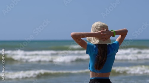 A teenage girl with a straw hat and glasses, with her hands folded behind her head, warms her face with the sun's rays and enjoys standing on the shore of the sea or ocean.
