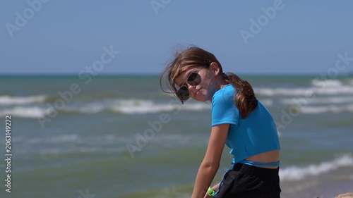 Young girl with long braided hair sitting alone on sandy beach surrounded by grass looking out over calm sea under cloudy sky