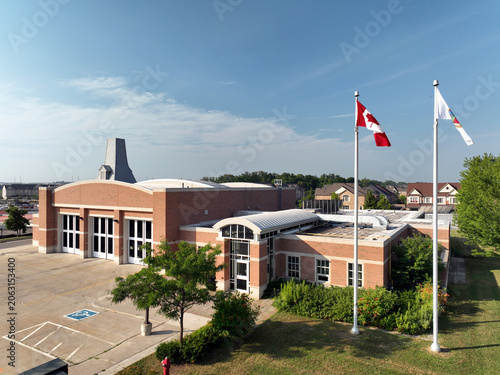 Fotografie Aerial view of a brick Canadian fire station house featuring large bay doors, arched entrance, Canadian flag, and residential neighbourhood backdrop on a sunny summer day
