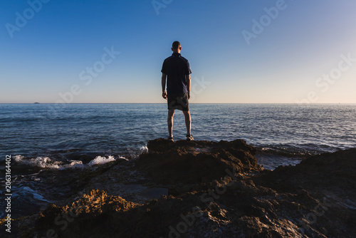 Rear view of a 43 year old man standing on dark rocks by the ocean, enjoying the scenic sea view during summer vacation in Crete, Greece