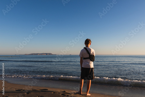 An 11 year old boy with a broken arm in a medical shoulder sling immobilizer bandage standing on a sandy beach looking at the sea during golden hour