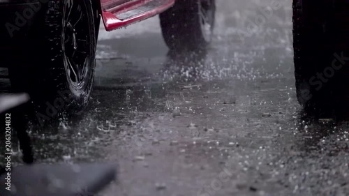 Rain drums on wet pavement beside red vehicle tires. No people, just water's rhythm against asphalt. Ideal for scenes of urban weather, driving, or moody atmosphere