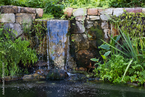 Stacked stone wall is channeling thin waterfall into pond, with moss, ferns, rippling surface