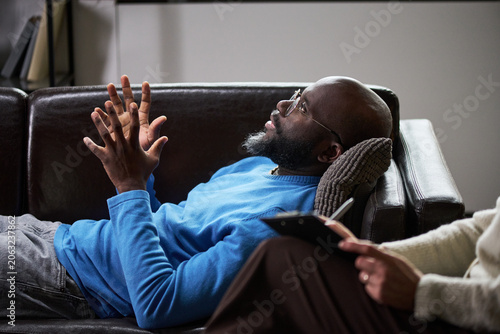 Photos Adult Black man reclining and speaking during therapy session while female therapist taking notes, conveying counseling and mental health support